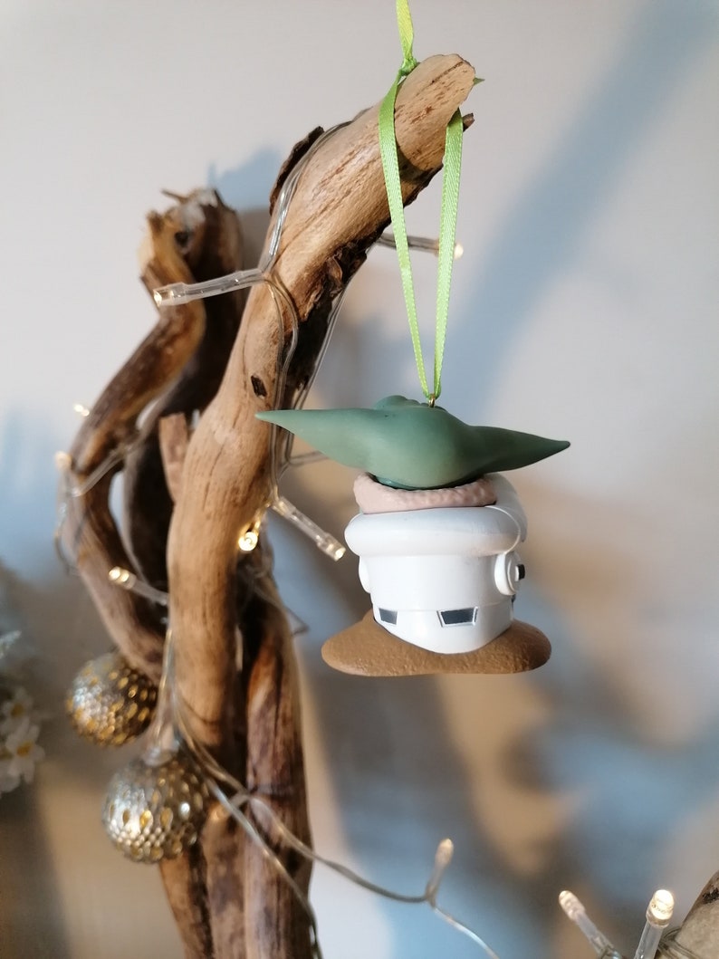 K&ouml;nnte beinhalten: Ein gr&uuml;ner und wei&szlig;er Weihnachtsbaumschmuck mit einer Figur aus dem Star Wars-Franchise. Der Schmuck h&auml;ngt an einem gr&uuml;nen Band an einem St&uuml;ck Treibholz.