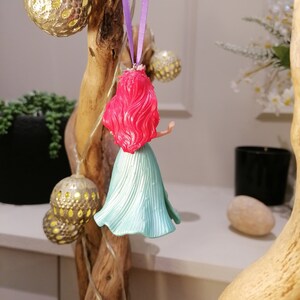 May include: A red-haired Ariel Disney character ornament with a teal blue dress hanging from a purple ribbon.