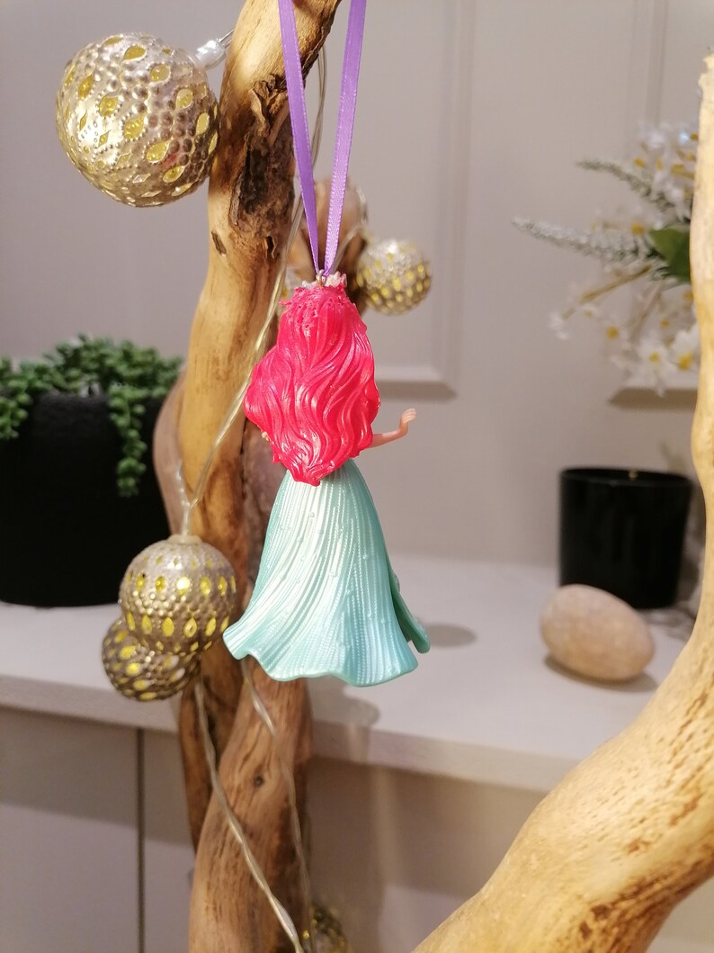 May include: A red-haired Ariel Disney character ornament with a teal blue dress hanging from a purple ribbon.