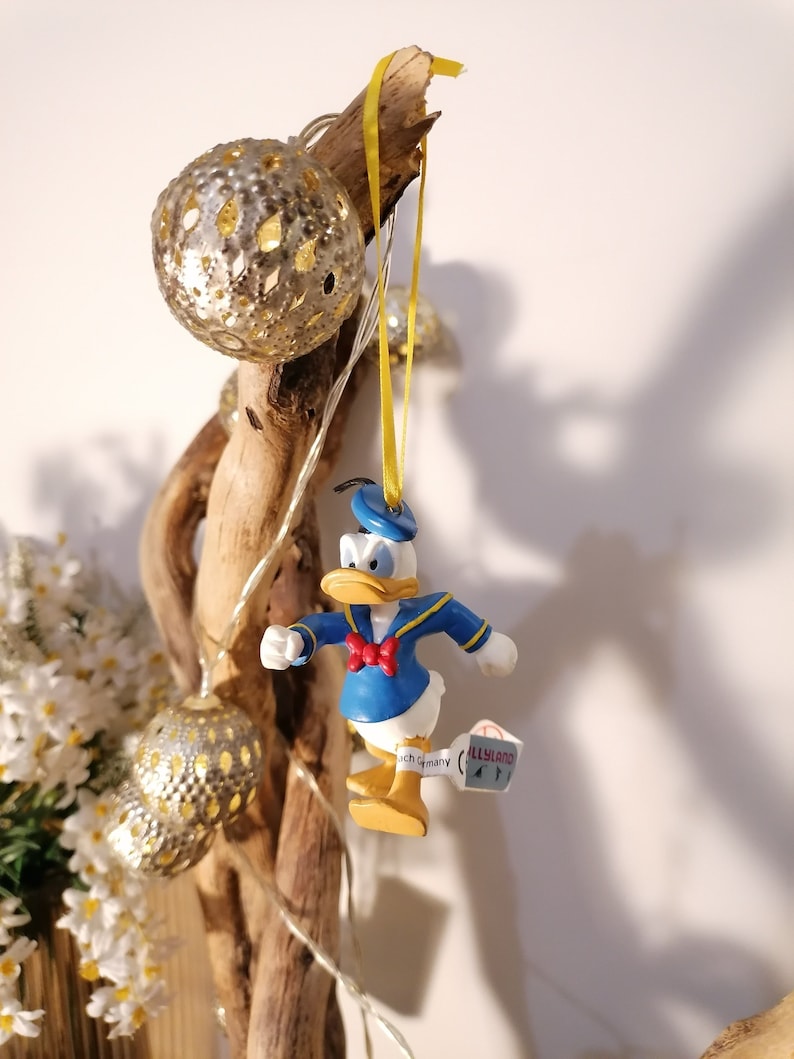以下が含まれることがあります： 水色と黄色のクリスマスオーナメントで、セーラー服を着たカモの漫画が描かれています。オーナメントは、ライトのついた枝にぶら下がっています。オーナメントには、「Allysland」と「Made in Germany」と書かれたタグが付いています。