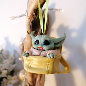 May include: A green and yellow Christmas ornament featuring a character from the Star Wars franchise, sitting in a yellow bag. The ornament is hanging from a green ribbon.