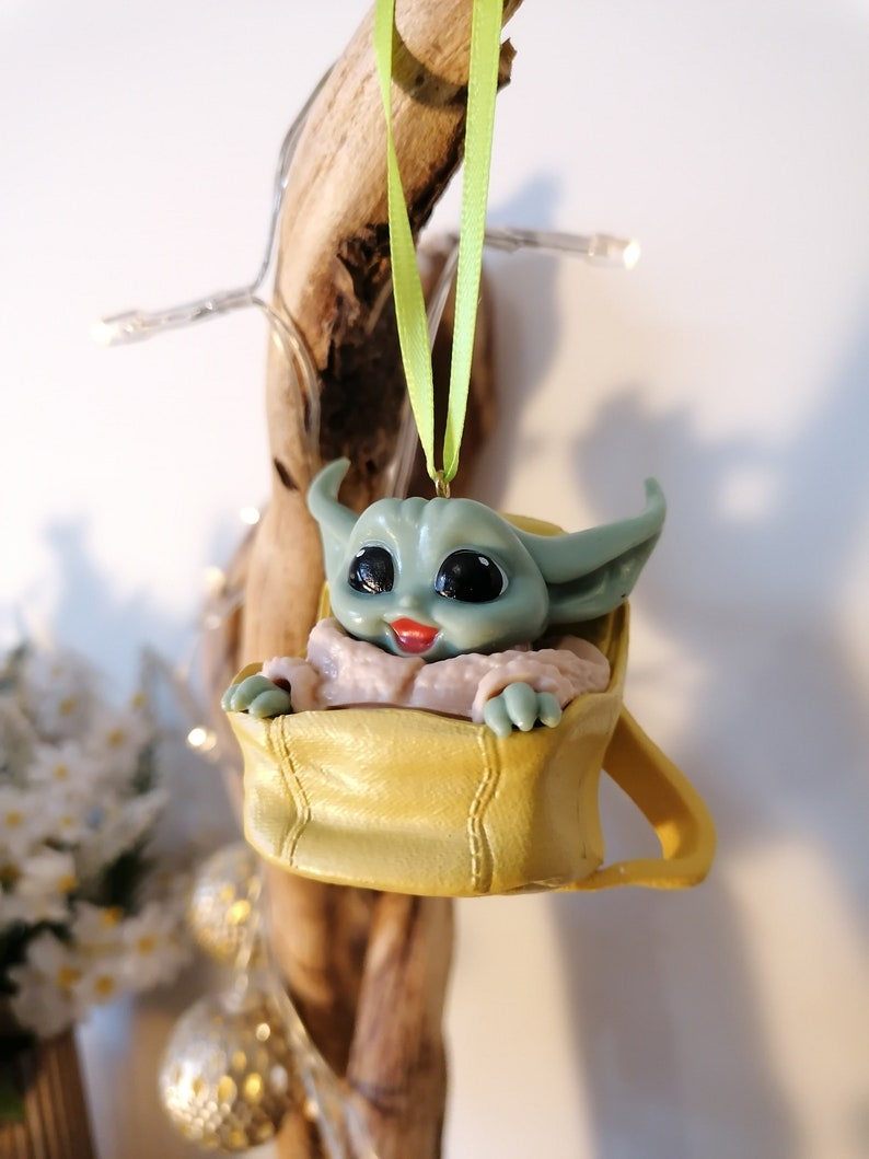 May include: A green and yellow Christmas ornament featuring a character from the Star Wars franchise, sitting in a yellow bag. The ornament is hanging from a green ribbon.
