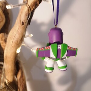 K&ouml;nnte beinhalten: Lila-gr&uuml;ner Weihnachtsbaumschmuck von Buzz Lightyear aus Toy Story. Der Schmuck h&auml;ngt an einem lila Band.