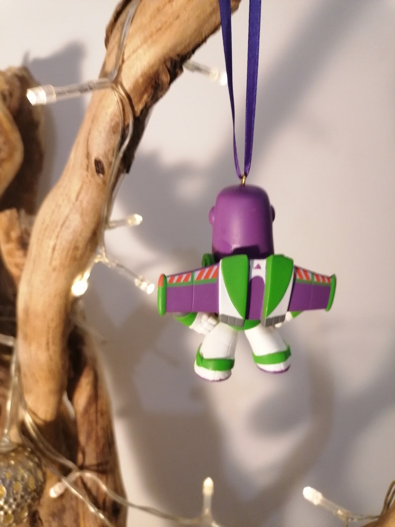 K&ouml;nnte beinhalten: Lila-gr&uuml;ner Weihnachtsbaumschmuck von Buzz Lightyear aus Toy Story. Der Schmuck h&auml;ngt an einem lila Band.