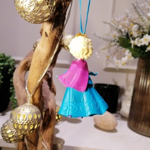 K&ouml;nnte beinhalten: Ein blauer und rosa Plastik-Puppen-Anh&auml;nger einer Prinzessin mit einer wei&szlig;en Krone und blonden Haaren. Die Puppe h&auml;ngt an einer blauen Schnur.