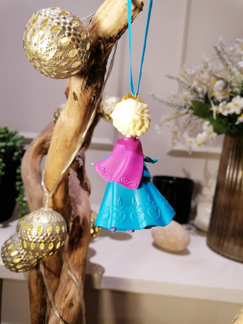 K&ouml;nnte beinhalten: Ein blauer und rosa Plastik-Puppen-Anh&auml;nger einer Prinzessin mit einer wei&szlig;en Krone und blonden Haaren. Die Puppe h&auml;ngt an einer blauen Schnur.