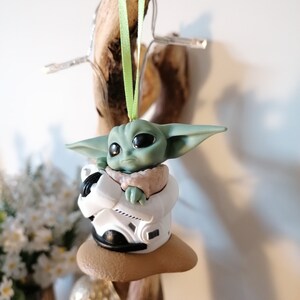 K&ouml;nnte beinhalten: Ein gr&uuml;ner und brauner Weihnachtsbaumschmuck mit einer Figur aus dem Star Wars-Franchise, The Mandalorian. Die Figur sitzt in einem kleinen, wei&szlig;en Raumschiff mit einer braunen Basis.