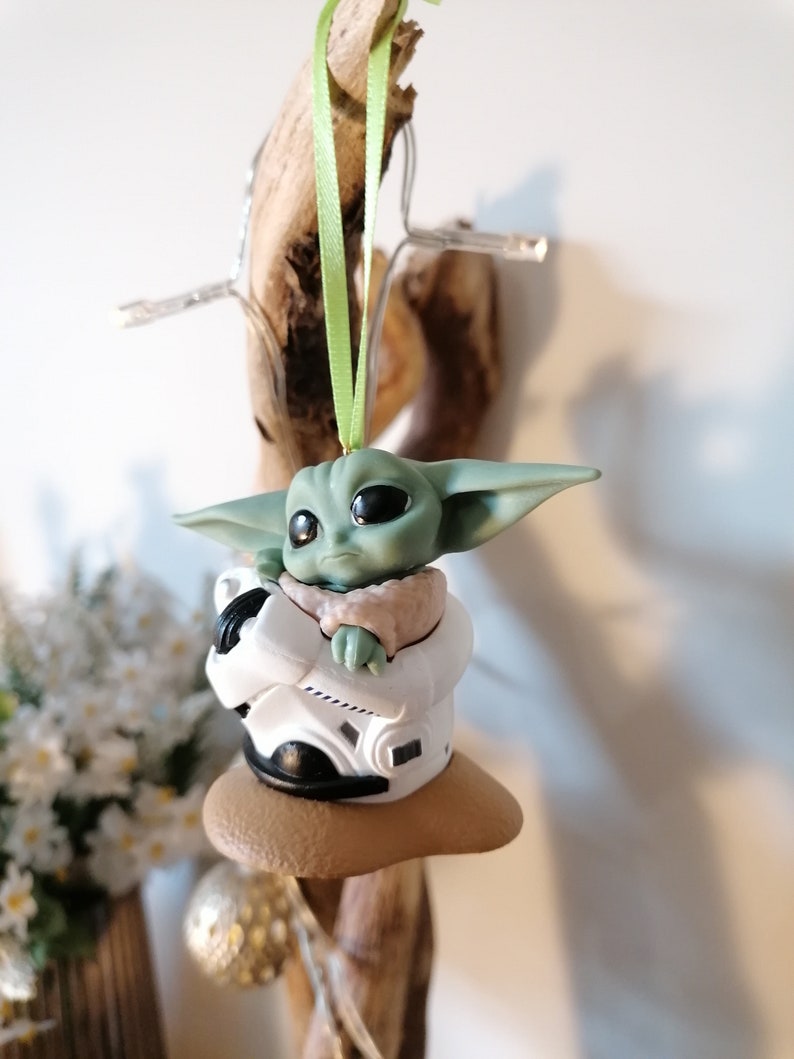 K&ouml;nnte beinhalten: Ein gr&uuml;ner und brauner Weihnachtsbaumschmuck mit einer Figur aus dem Star Wars-Franchise, The Mandalorian. Die Figur sitzt in einem kleinen, wei&szlig;en Raumschiff mit einer braunen Basis.