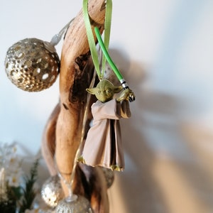 Puede incluir: Adorno navide&ntilde;o de Yoda con un sable de luz verde y una t&uacute;nica beige, suspendido de una cinta verde. La figura de Yoda est&aacute; unida a un trozo de madera flotante. Tambi&eacute;n se ven adornos de globo plateados y luces de hadas.