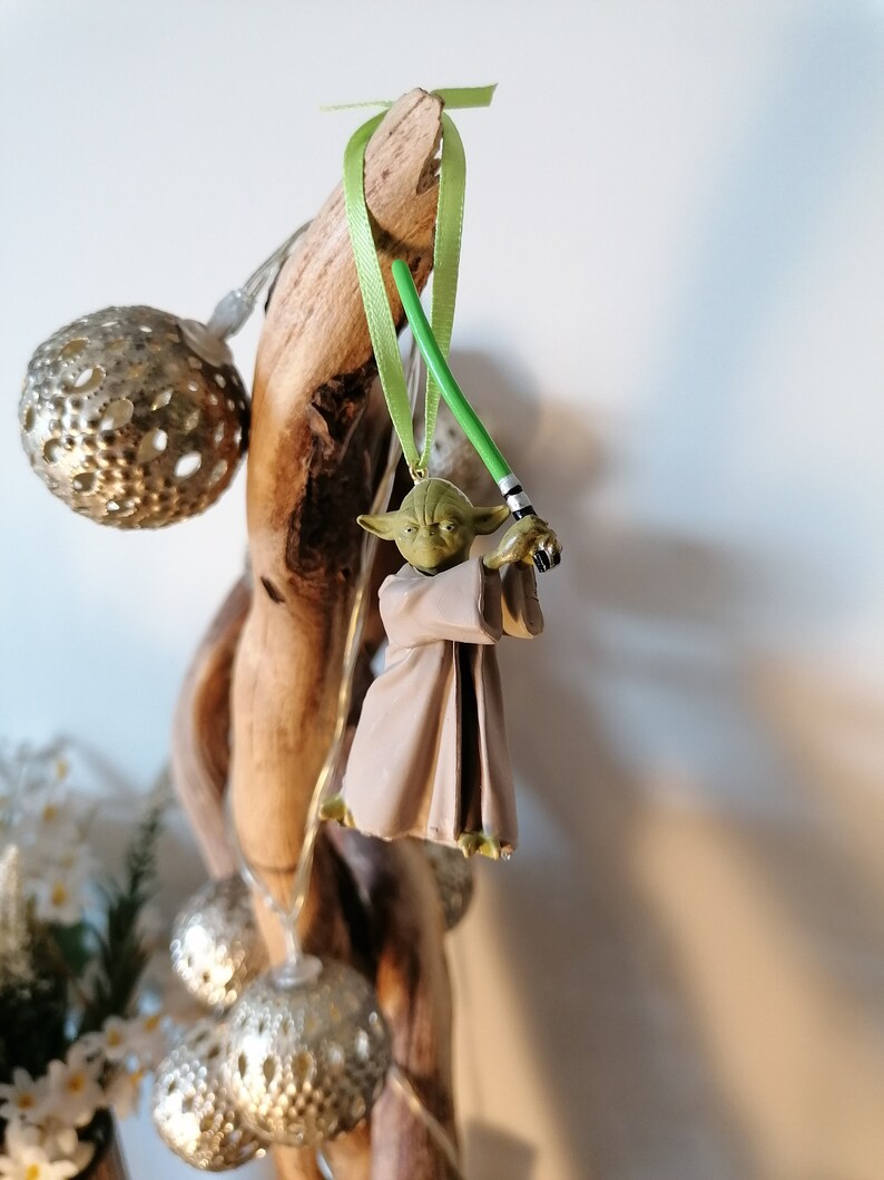 Puede incluir: Adorno navide&ntilde;o de Yoda con un sable de luz verde y una t&uacute;nica beige, suspendido de una cinta verde. La figura de Yoda est&aacute; unida a un trozo de madera flotante. Tambi&eacute;n se ven adornos de globo plateados y luces de hadas.