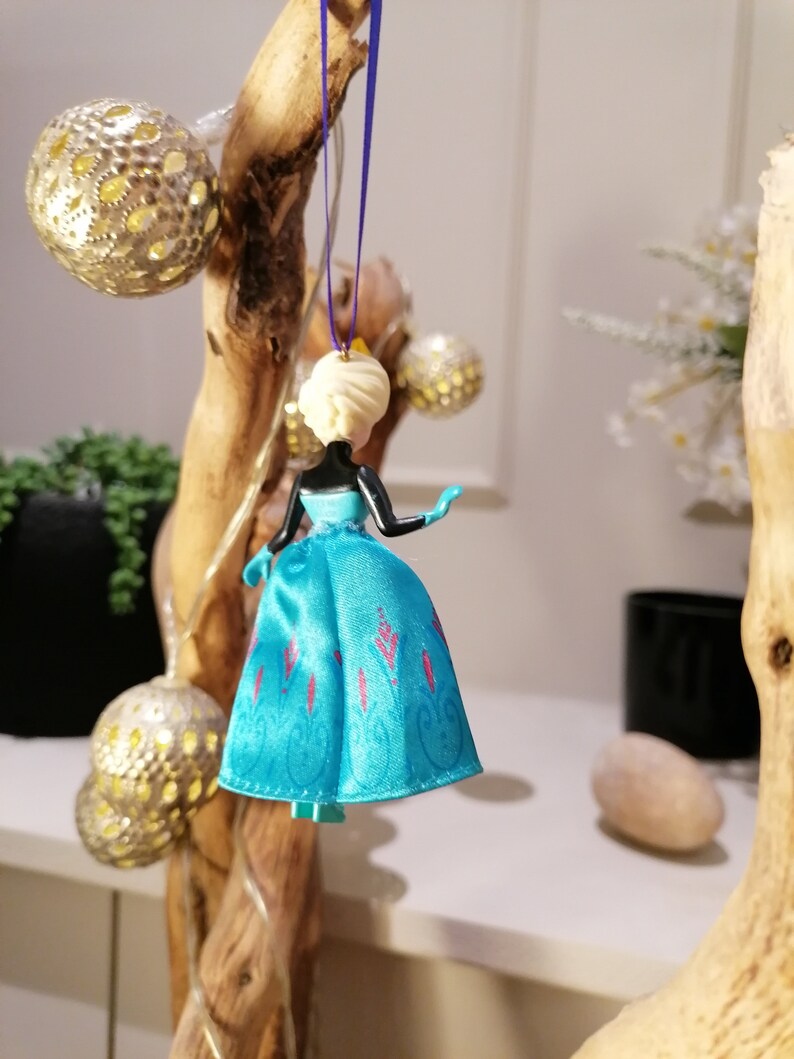K&ouml;nnte beinhalten: Ein blauer und rosa Plastik-Puppen-Anh&auml;nger einer Prinzessin, die ein blaues Kleid mit rosa floralen Akzenten tr&auml;gt. Die Puppe h&auml;ngt an einer lila Schnur.