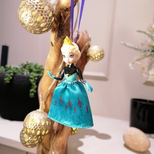 K&ouml;nnte beinhalten: Ein blauer und wei&szlig;er Plastik-Puppen-Anh&auml;nger einer Prinzessin, die ein blaues Kleid mit roten und wei&szlig;en Akzenten tr&auml;gt. Die Puppe h&auml;ngt an einem lila Band an einem Holzast.