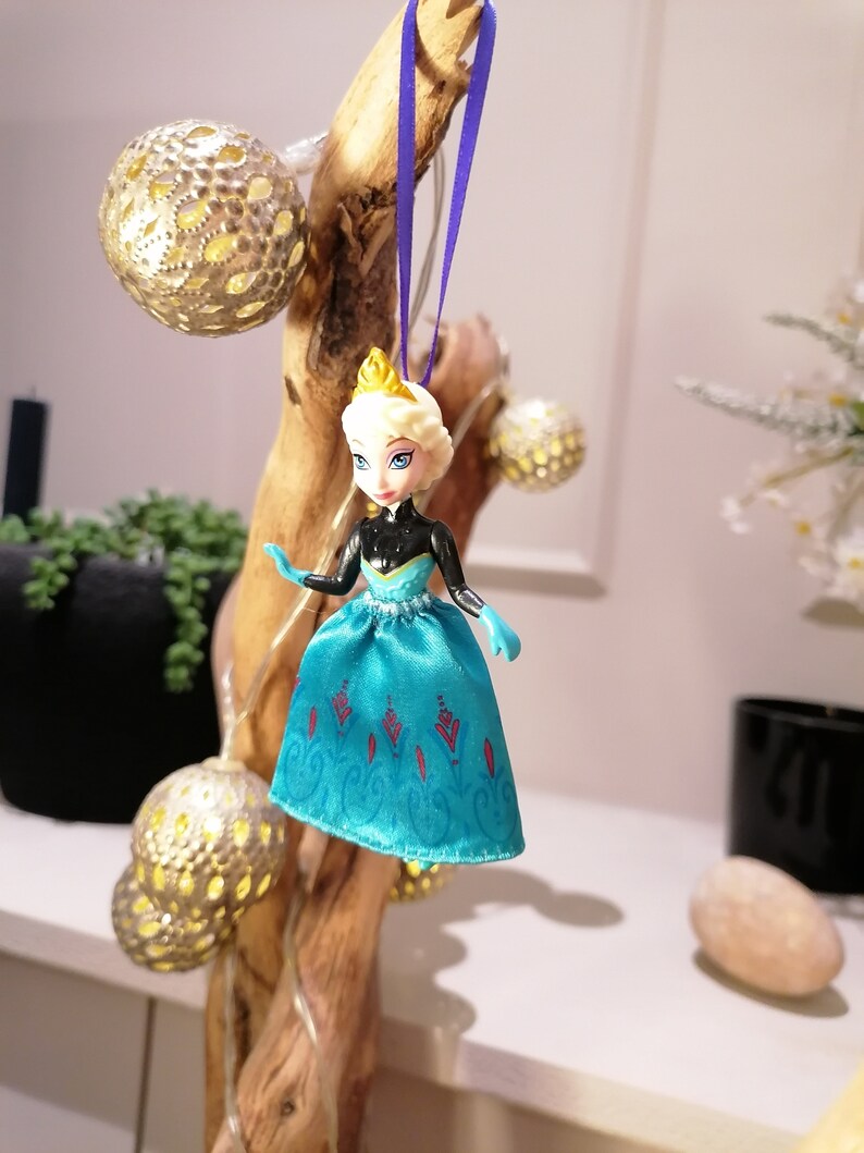 K&ouml;nnte beinhalten: Ein blauer und wei&szlig;er Plastik-Puppen-Anh&auml;nger einer Prinzessin, die ein blaues Kleid mit roten und wei&szlig;en Akzenten tr&auml;gt. Die Puppe h&auml;ngt an einem lila Band an einem Holzast.