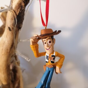 May include: A red string hangs from the top of the image, with a plastic toy figurine of Woody from Toy Story hanging from the string. Woody is wearing his signature brown cowboy hat, a yellow and brown plaid shirt, a blue vest with a sheriff's star, and blue jeans. He is holding his hat in his left hand and smiling.