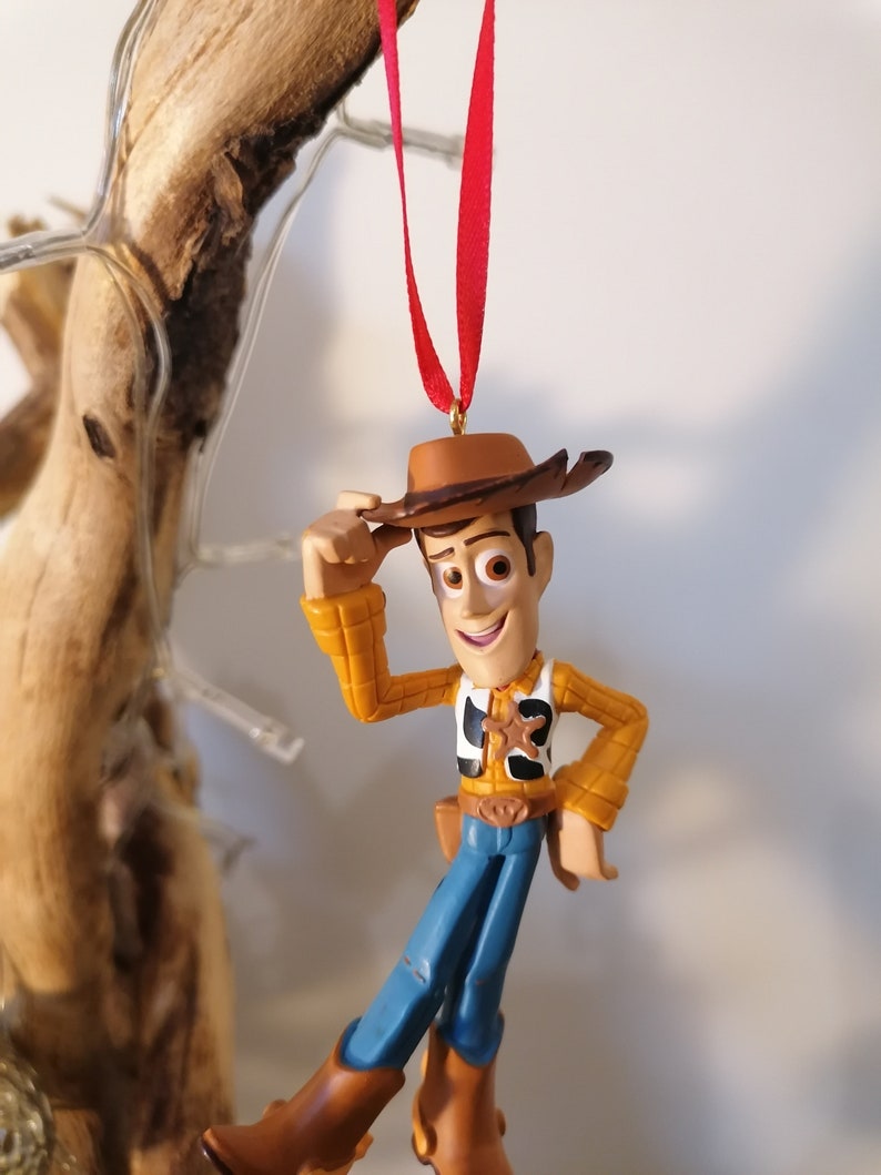 May include: A red string hangs from the top of the image, with a plastic toy figurine of Woody from Toy Story hanging from the string. Woody is wearing his signature brown cowboy hat, a yellow and brown plaid shirt, a blue vest with a sheriff's star, and blue jeans. He is holding his hat in his left hand and smiling.