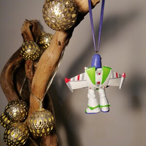 Pu&ograve; includere: Un ornamento di plastica bianco, verde e blu di Buzz Lightyear della serie di film Toy Story. L'ornamento &egrave; appeso a un nastro viola su un ramo con palline decorative dorate.