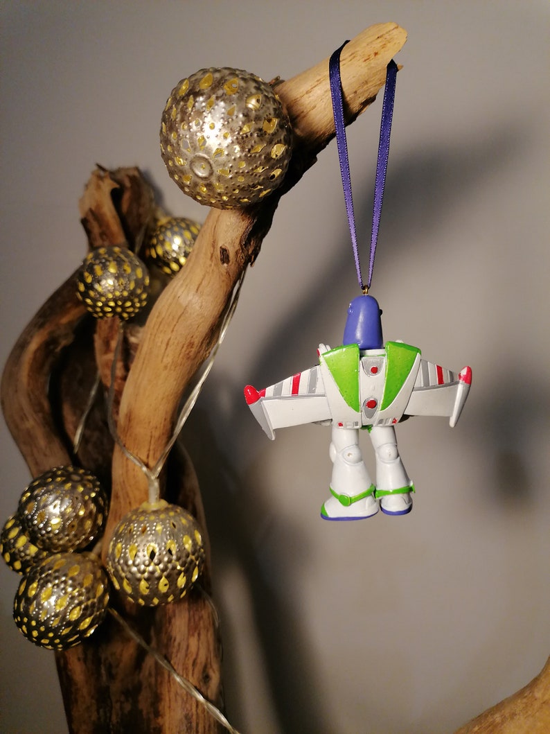 Pu&ograve; includere: Un ornamento di plastica bianco, verde e blu di Buzz Lightyear della serie di film Toy Story. L'ornamento &egrave; appeso a un nastro viola su un ramo con palline decorative dorate.