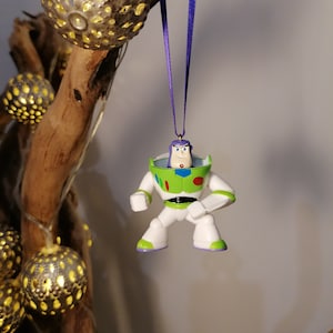 Puede incluir: Un adorno de plástico verde, blanco y morado de Buzz Lightyear de la franquicia Toy Story. El adorno está colgado de una cinta morada.