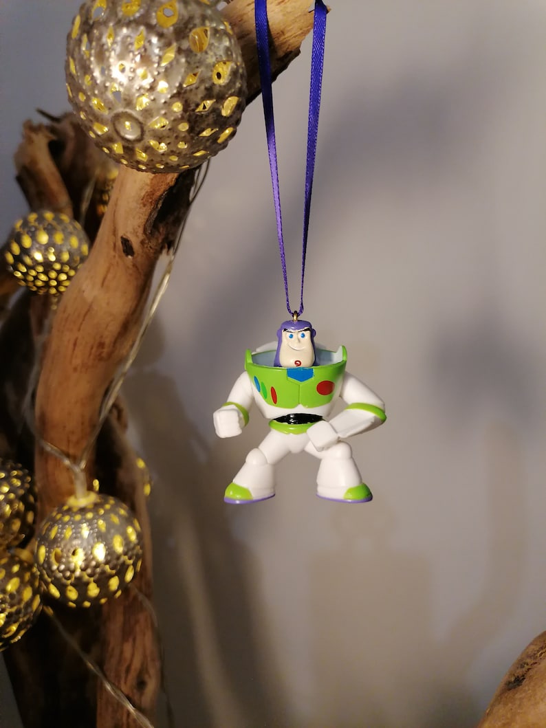May include: A green, white and purple plastic ornament of Buzz Lightyear from the Toy Story franchise. The ornament is hanging from a purple ribbon.