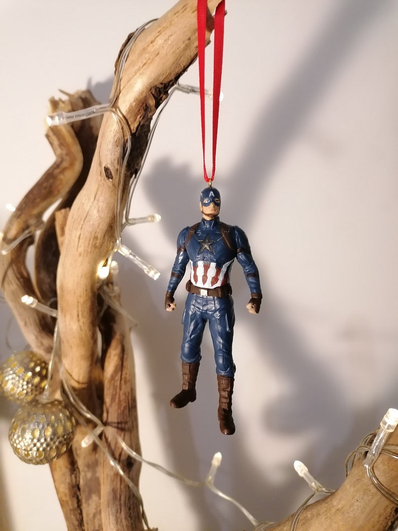 May include: A red string hangs from a piece of driftwood with a Captain America Christmas ornament attached. The ornament is a plastic figurine of Captain America in his blue and red suit with a white star on his chest.