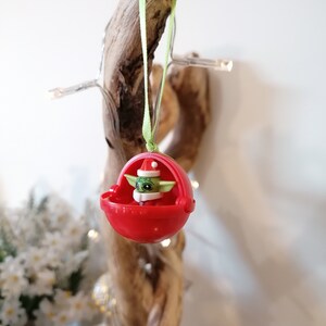 Pu&ograve; includere: Un ornamento natalizio rosso con un personaggio verde che indossa un cappello da Babbo Natale. L'ornamento &egrave; appeso a un nastro verde su un pezzo di legno di deriva.