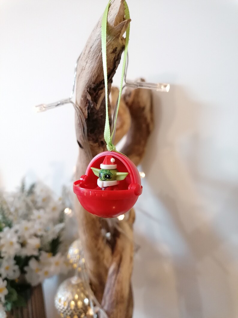 Pu&ograve; includere: Un ornamento natalizio rosso con un personaggio verde che indossa un cappello da Babbo Natale. L'ornamento &egrave; appeso a un nastro verde su un pezzo di legno di deriva.