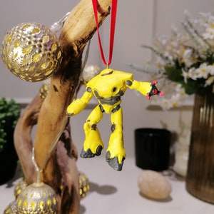 Puede incluir: Adorno de robot amarillo con detalles negros colgando de una cinta roja en una rama con luces doradas y blancas.