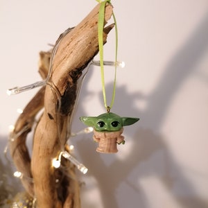 May include: A green and tan Baby Yoda ornament hanging from a light green ribbon. The ornament is attached to a piece of driftwood. Fairy lights are wrapped around the driftwood.
