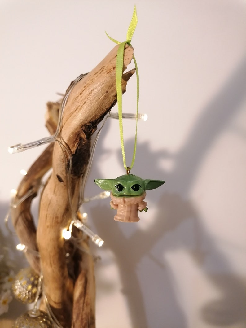 May include: A green and tan Baby Yoda ornament hanging from a light green ribbon. The ornament is attached to a piece of driftwood. Fairy lights are wrapped around the driftwood.