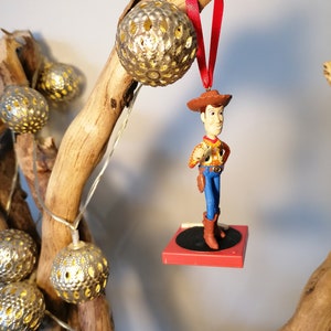 Puede incluir: Un adorno navide&ntilde;o rojo y blanco con un personaje de vaquero de juguete de pie sobre un tocadiscos rojo y negro. El adorno est&aacute; colgado de una rama con luces de cadena plateadas y blancas.