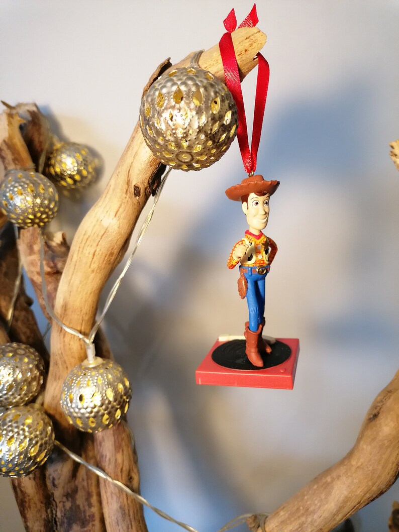 Puede incluir: Un adorno navide&ntilde;o rojo y blanco con un personaje de vaquero de juguete de pie sobre un tocadiscos rojo y negro. El adorno est&aacute; colgado de una rama con luces de cadena plateadas y blancas.