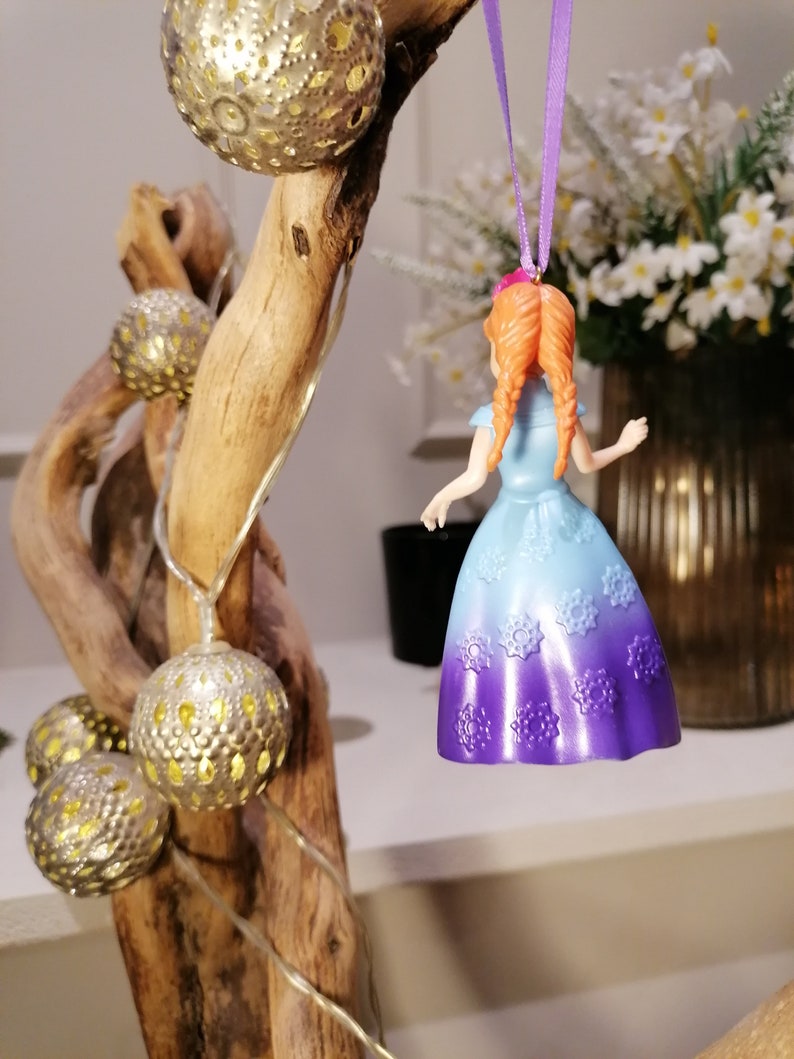 K&ouml;nnte beinhalten: Ein lila und blauer Prinzessinnen-Anh&auml;nger mit einem wei&szlig;en Schneeflockenmuster, der an einem lila Band h&auml;ngt. Der Anh&auml;nger ist aus Kunststoff und hat ein detailliertes Design.