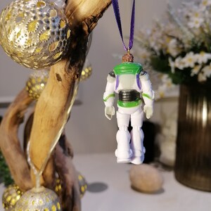 May include: A white and green plastic ornament of Buzz Lightyear from the Toy Story franchise. The ornament is hanging from a purple ribbon and is facing away from the camera.