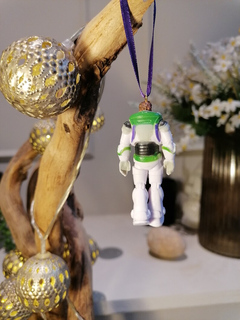 May include: A white and green plastic ornament of Buzz Lightyear from the Toy Story franchise. The ornament is hanging from a purple ribbon and is facing away from the camera.