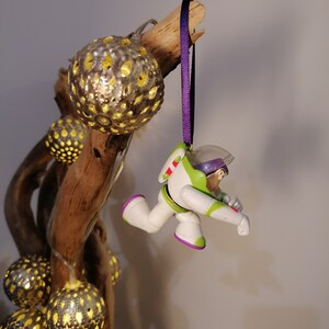 Puede incluir: Un adorno navide&ntilde;o de pl&aacute;stico morado y blanco con una figura de Buzz Lightyear de la franquicia Toy Story. El adorno est&aacute; colgado de una cinta morada en una rama con luces decorativas.
