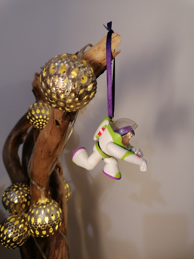 Puede incluir: Un adorno navide&ntilde;o de pl&aacute;stico morado y blanco con una figura de Buzz Lightyear de la franquicia Toy Story. El adorno est&aacute; colgado de una cinta morada en una rama con luces decorativas.