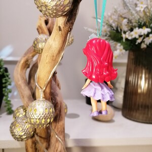 Peut inclure: Une poup&eacute;e rouge &agrave; cheveux, v&ecirc;tue d'une robe violette, est suspendue &agrave; une branche avec un ruban turquoise. La poup&eacute;e porte une robe violette avec une bordure blanche et un n&oelig;ud rouge dans les cheveux. La branche est d&eacute;cor&eacute;e d'ornements dor&eacute;s et blancs.