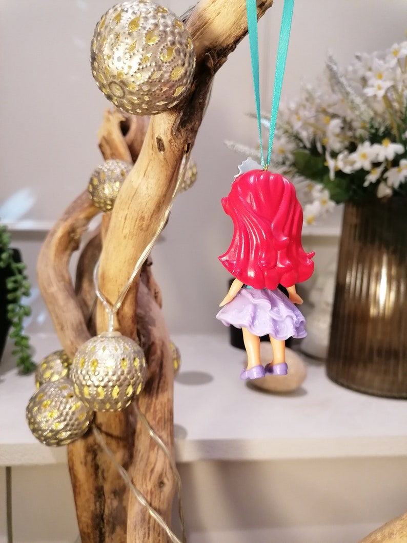 Peut inclure: Une poup&eacute;e rouge &agrave; cheveux, v&ecirc;tue d'une robe violette, est suspendue &agrave; une branche avec un ruban turquoise. La poup&eacute;e porte une robe violette avec une bordure blanche et un n&oelig;ud rouge dans les cheveux. La branche est d&eacute;cor&eacute;e d'ornements dor&eacute;s et blancs.