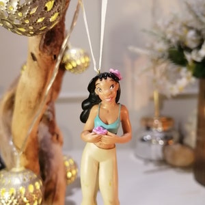 May include: A brown-haired figurine of a woman wearing a blue tank top and white pants. She is holding a pink flower and has a pink flower in her hair. The figurine is hanging from a white string.
