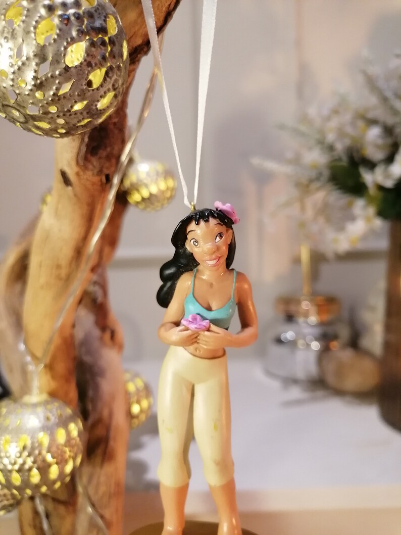 May include: A brown-haired figurine of a woman wearing a blue tank top and white pants. She is holding a pink flower and has a pink flower in her hair. The figurine is hanging from a white string.