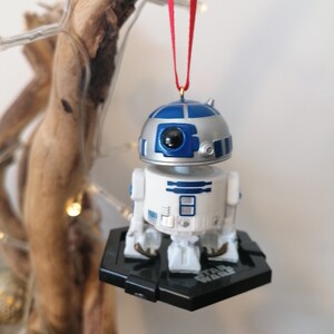 May include: A white and blue R2-D2 Star Wars character ornament hanging from a red ribbon. The ornament is attached to a black base with the words "Star Wars" printed on it.