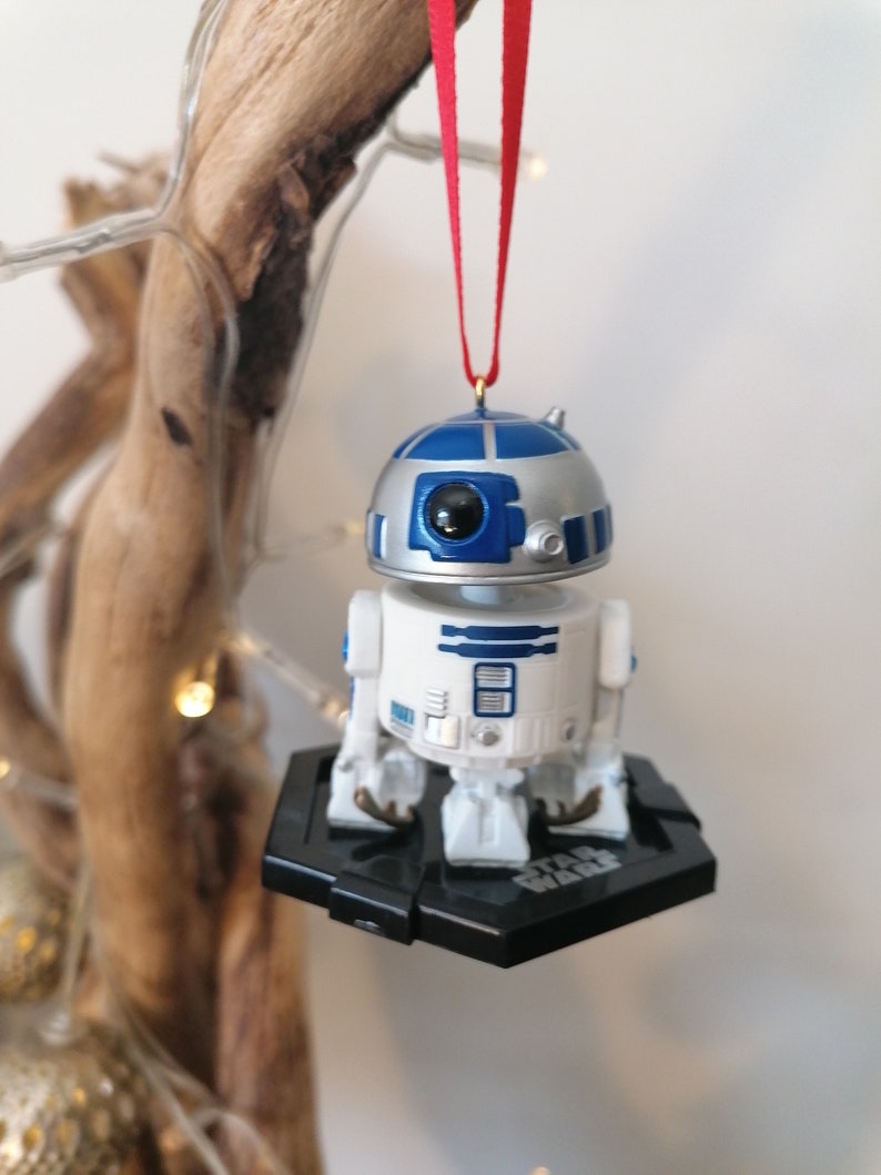 May include: A white and blue R2-D2 Star Wars character ornament hanging from a red ribbon. The ornament is attached to a black base with the words "Star Wars" printed on it.