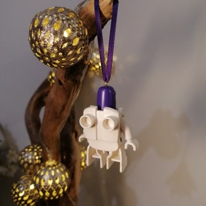 May include: White Lego figure with a purple head hanging from a purple ribbon on a branch with gold and white string lights.
