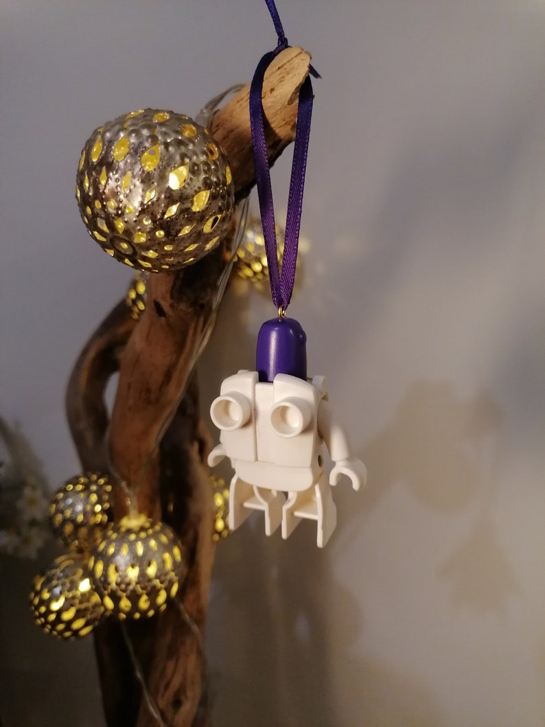 May include: White Lego figure with a purple head hanging from a purple ribbon on a branch with gold and white string lights.