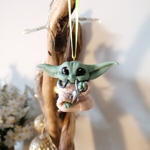 May include: A green and pink Yoda ornament with a silver staff hanging from a green ribbon. The ornament is made of polymer clay and is a handmade creation.