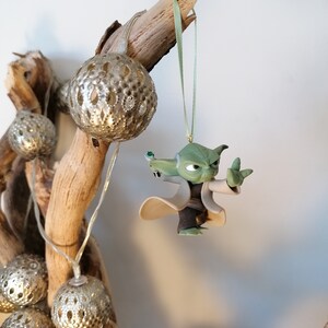 May include: A green Yoda character ornament hanging from a branch with silver and white decorative balls. The ornament is made of plastic and has a green, brown, and white color scheme.