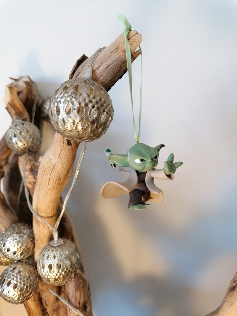 May include: A green Yoda character ornament hanging from a branch with silver and white decorative balls. The ornament is made of plastic and has a green, brown, and white color scheme.