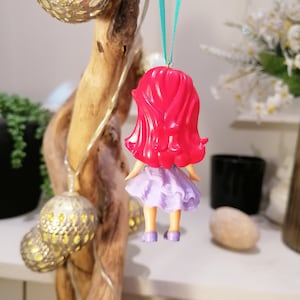 Peut inclure: Une poup&eacute;e aux cheveux roux portant une robe violette et des chaussures bleues est suspendue &agrave; un ruban vert. La poup&eacute;e est une d&eacute;coration de No&euml;l.