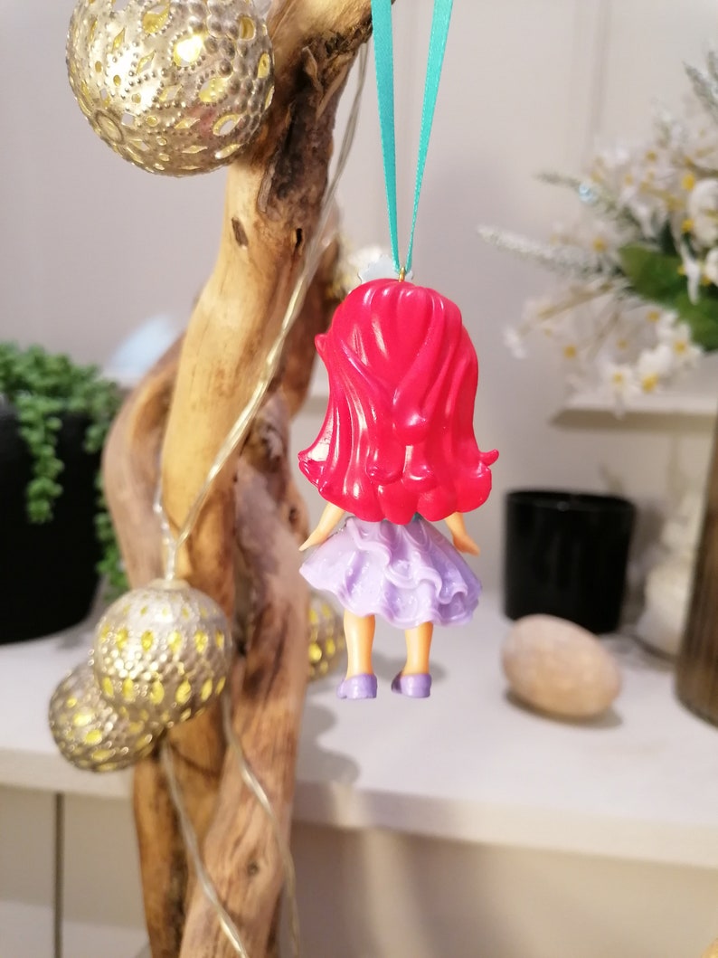 Peut inclure: Une poup&eacute;e aux cheveux roux portant une robe violette et des chaussures bleues est suspendue &agrave; un ruban vert. La poup&eacute;e est une d&eacute;coration de No&euml;l.