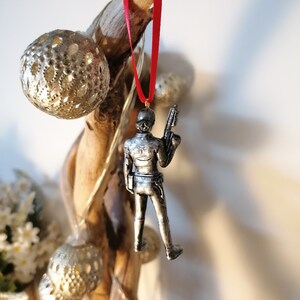 May include: A silver-coloured figurine of a person holding a weapon, hanging from a red ribbon on a branch. The figurine is a Christmas ornament.